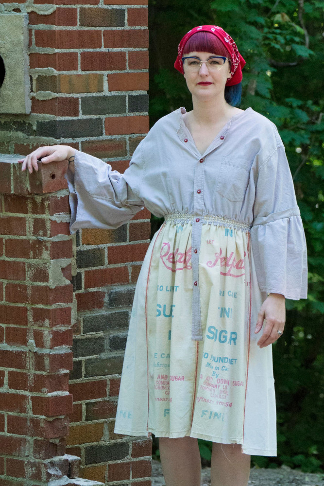 Model stands beside old outdoor fireplace. She is wearing a dress made from a man's cotton button down shirt and several sugar bags.