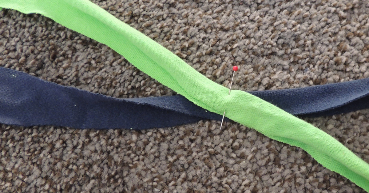 A green strip of t-shirt yarn is laid across a blue strip and they are pinned together where they cross.  Both strips were folded in half lengthwise before they were put in position.